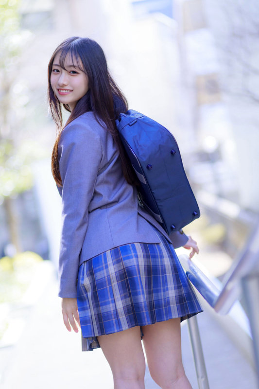 長い黒髪の女性がグレーのブレザーと青チェックのスカート姿で、紺色のリュックを背負い屋外の手すりを持って振り返る