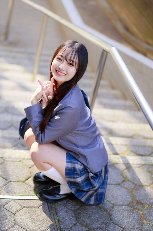 グレーのブレザーに青チェックのスカート姿の女性が、屋外の階段にしゃがみ両手でピースをして微笑む