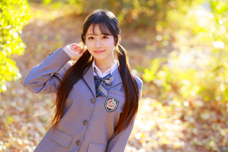 黄葉の屋外で、グレーのブレザー制服にチェックのリボンをつけた長い髪の女性が髪に手を添えて微笑んでいる