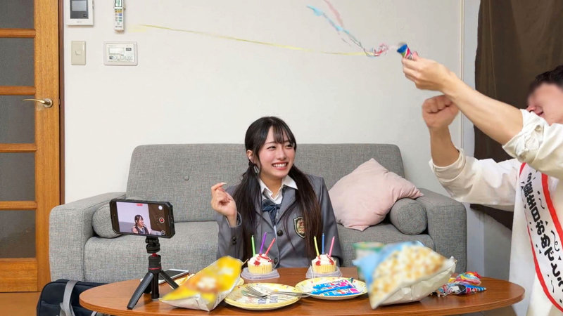 リビングのソファに制服で座る女性が笑い、テーブルのカップケーキの前で男性がクラッカーを鳴らしている