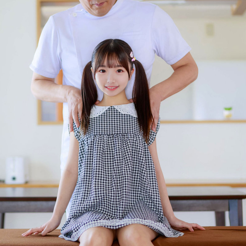 ギンガムチェックのワンピース姿でマッサージ台に腰掛け、背後から男性に肩を触れられながらこちらを見つめるカナちゃんのサンプル画像
